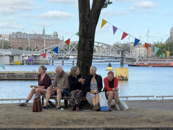 Five people are seen relaxing on a bench under a tree by the water at Commandantsbrug, Marineterrein, Amsterdam